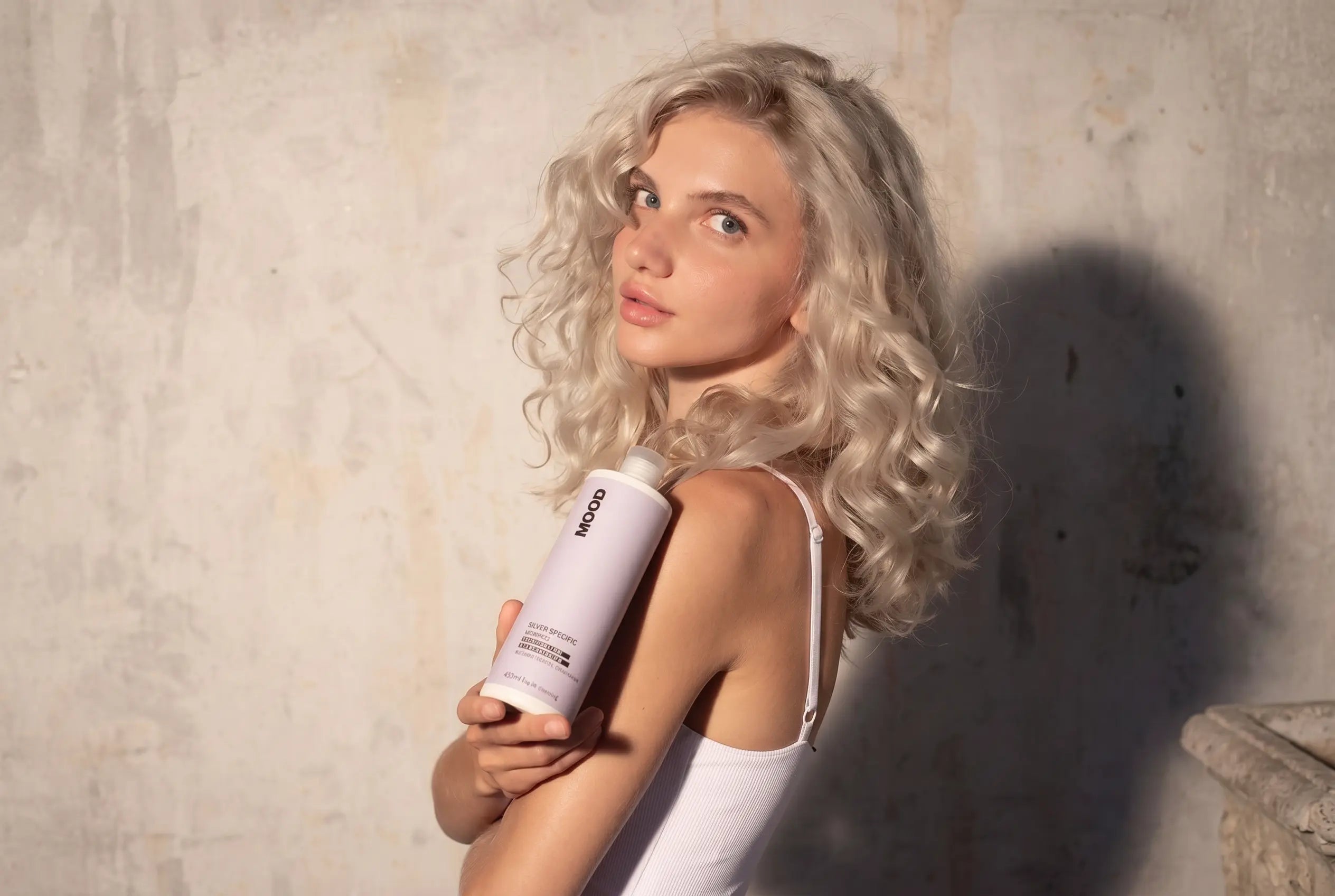 Model met blond, golvend haar die een Silver Specific haarverzorgingsproduct vasthoudt. Ideaal voor blond, wit, grijs of gehighlight haar om ongewenste gele tinten te neutraliseren en het haar meer glans te geven.